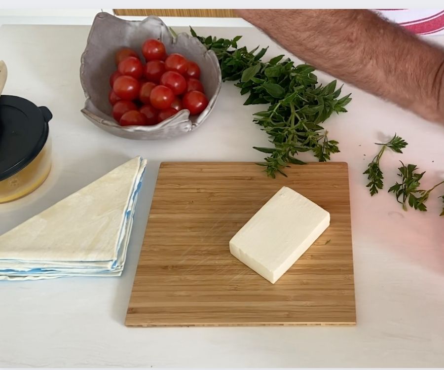 A board with a block of feta, a bowl of cherry tomatoes, pastry triangles, melted butter, fresh herbs